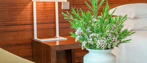 Habitación de hotel con cabecera de madera, mesita de noche con lámpara blanca y un florero con follaje verde y flores blancas.