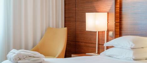 A hotel room with a bed, a bedside table with a lamp, a chair, and a rolled-up towel.