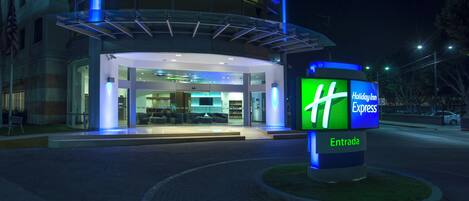 Edificio de Holiday Inn Express con entrada de vidrio, un cartel que indica la entrada y un interior bien iluminado.