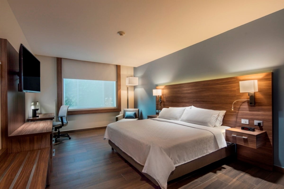 A modern hotel room with a large bed, a desk, a chair, a TV, and a window.