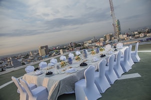 Una larga mesa de comedor dispuesta para un evento formal con sillas blancas, una vista de la ciudad al fondo y una grúa visible a lo lejos.