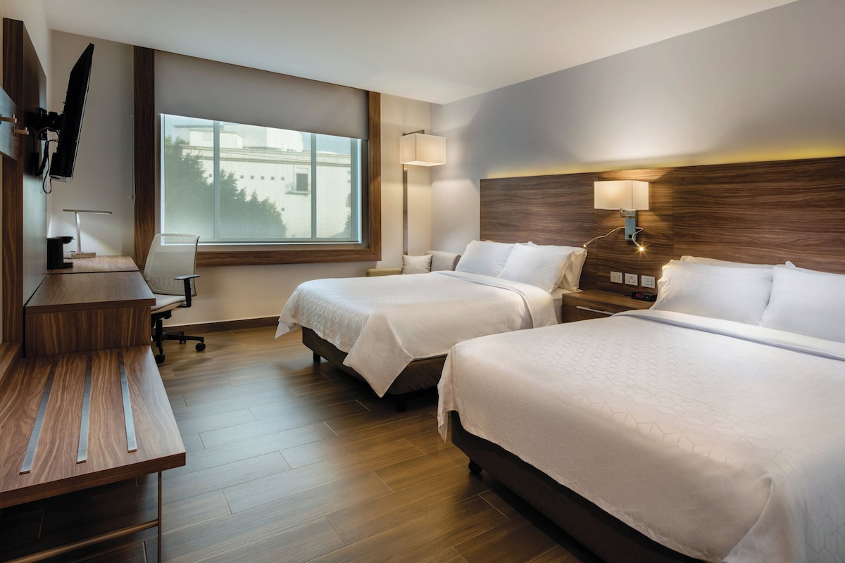 A hotel room with two beds, a desk, a chair, a TV, and a window.