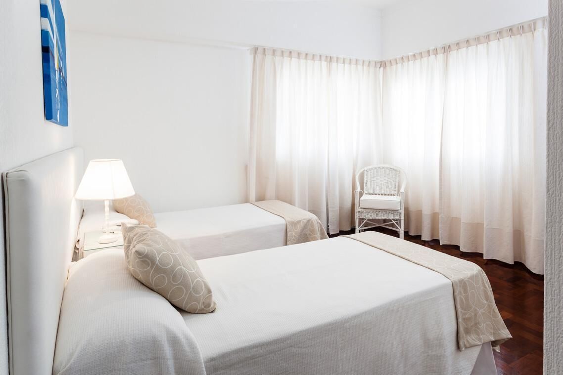 Habitació d'hotel amb dos llits, una nit falda amb una làmpada, una cadira i cortines translúcides.