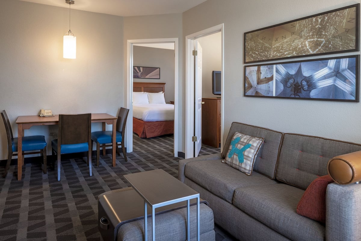 A hotel room with a bed, a sofa, a desk with chairs, a television, and artwork on the wall.