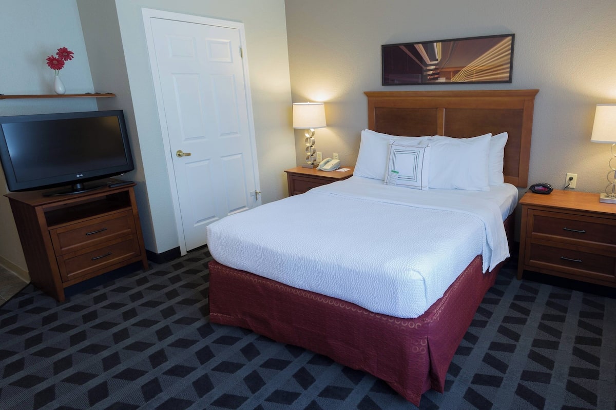 A hotel room with a large bed, a television on a wooden stand, a bedside table with a lamp, and a door.