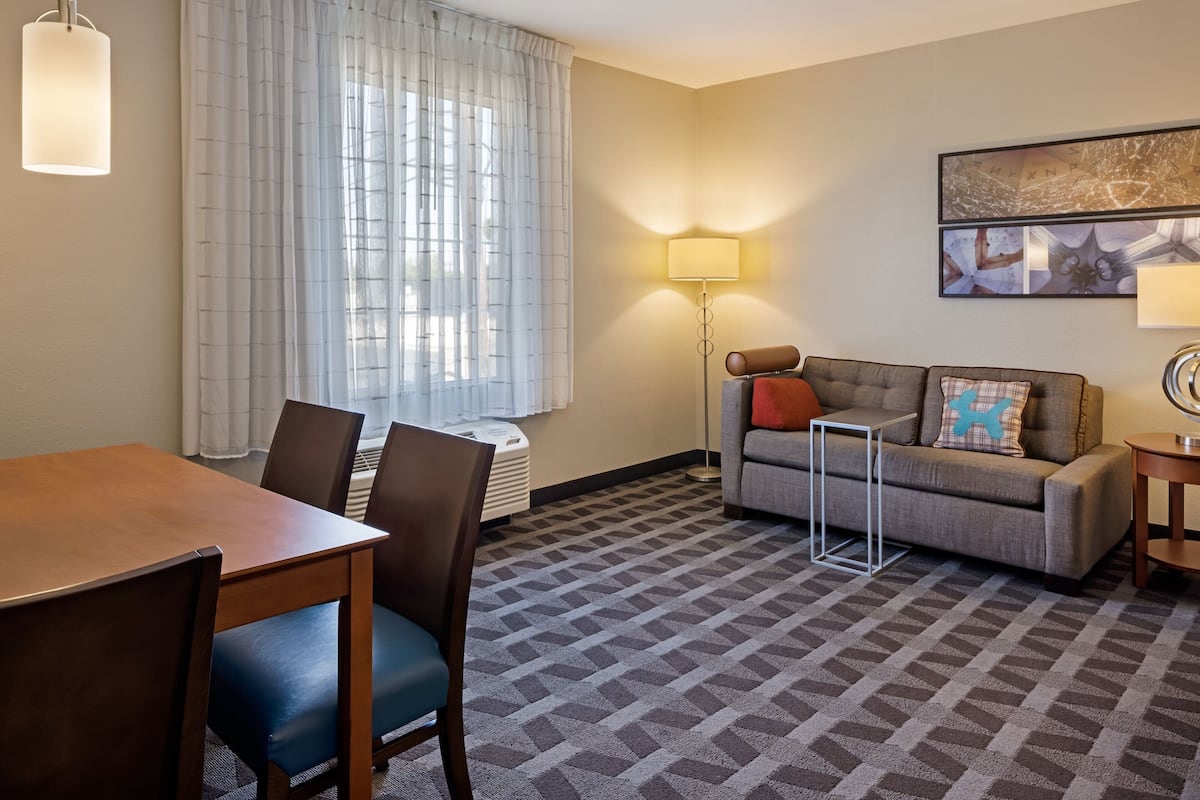A hotel room with a dining table, chairs, a sofa, a lamp, and a framed artwork on the wall.