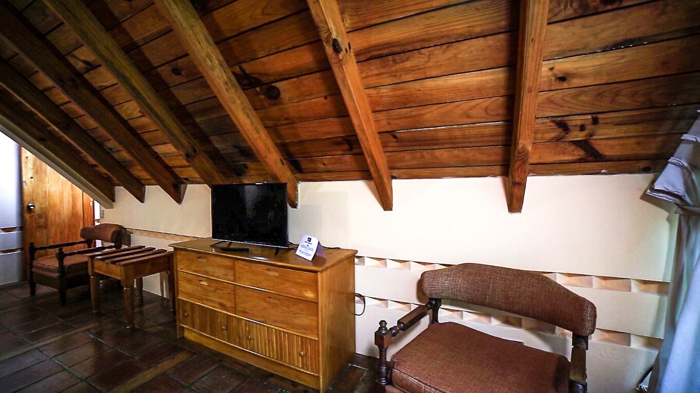 A rustic wooden ceiling with exposed beams, a wooden dresser with a television on top, a cushioned chair, and a small table.