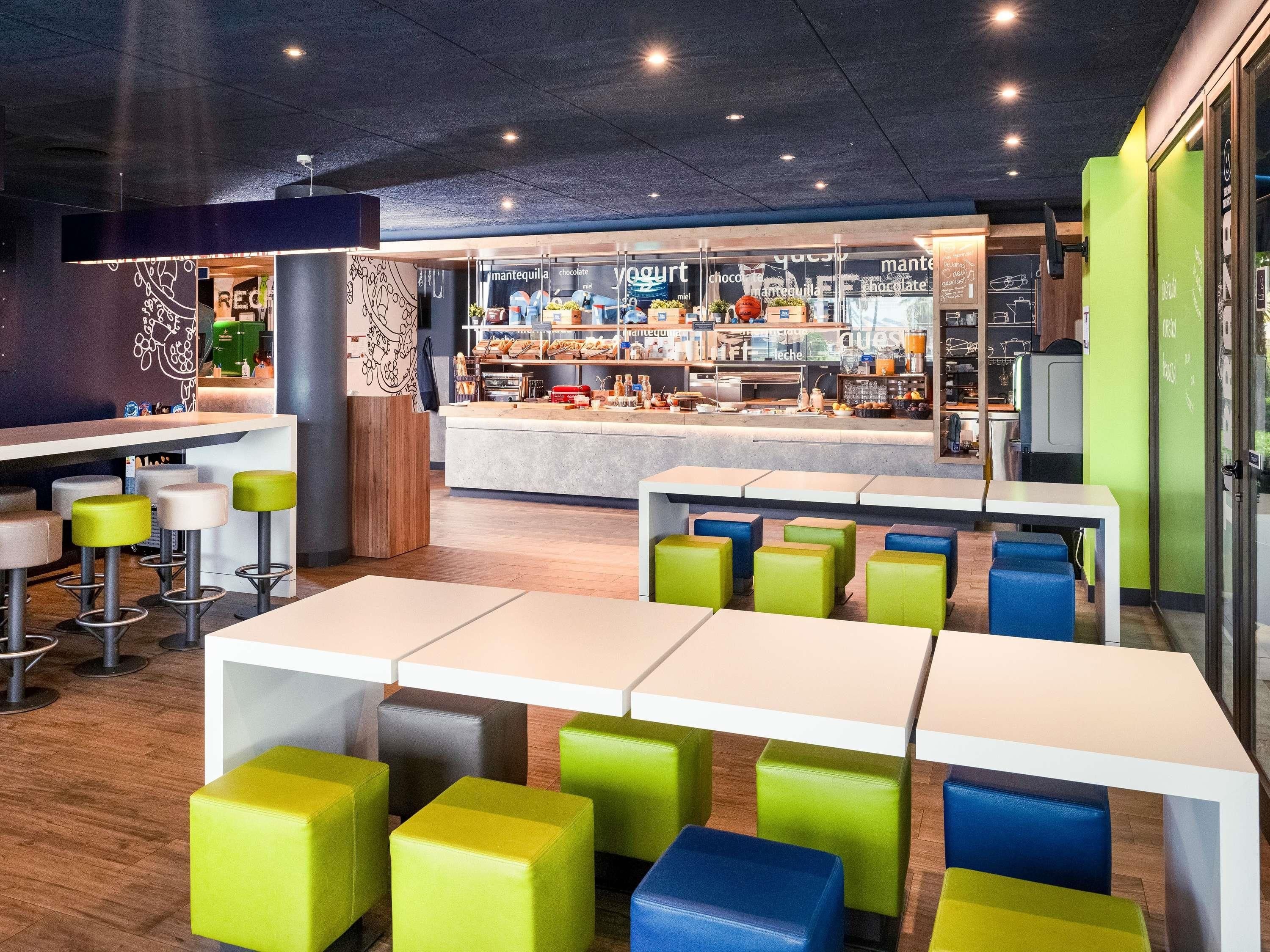 Interior de una cafetería moderna con varias zonas de estar, una barra con diferentes opciones de comida y una cocina a la vista.