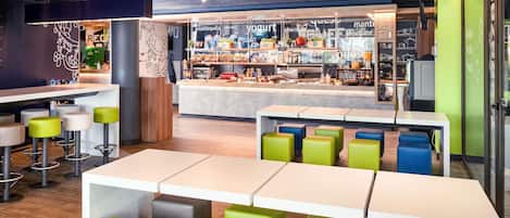 Interior de una cafetería moderna con varias zonas de estar, una barra con diferentes opciones de comida y una cocina a la vista.
