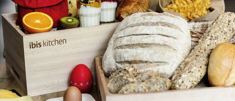 A wooden box labeled "ibis kitchen" containing various breads, a croissant, and a red egg with a timer.