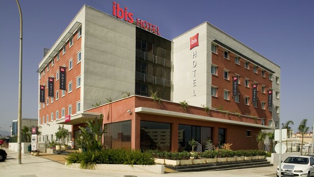 Un hotel moderno con fachada roja y gris, varios balcones y un llamativo letrero de 'ibis Hotel'.