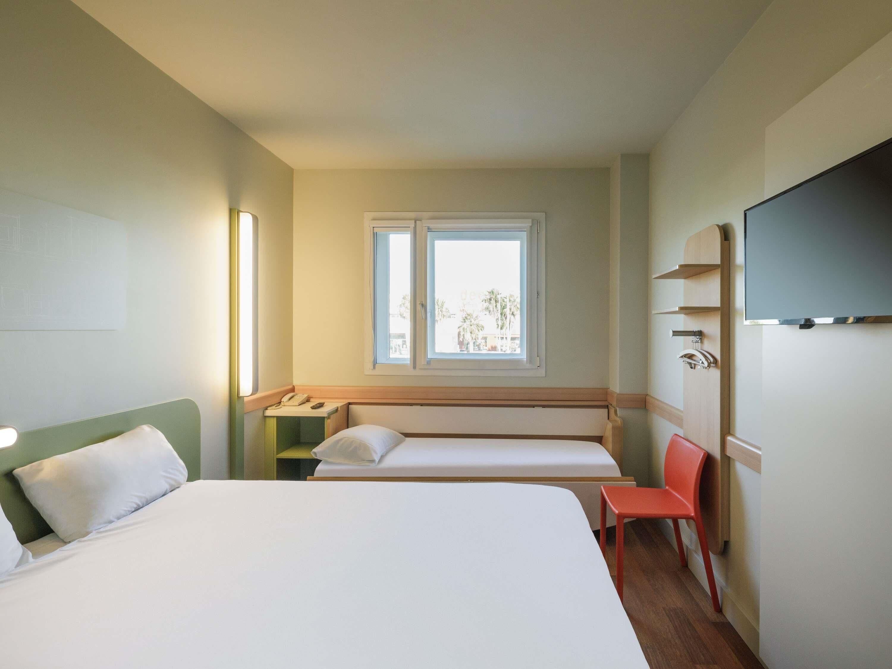 A hotel room with a large bed, a smaller bed, a desk with a phone, a chair, a TV, and a window with a view.