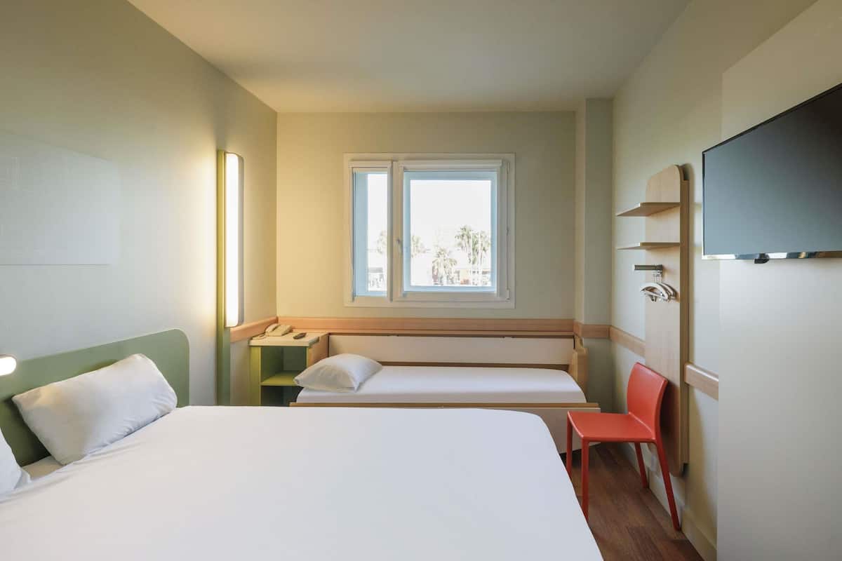 A hotel room with a large bed, a smaller bed, a desk with a phone, a chair, a TV, and a window with a view.