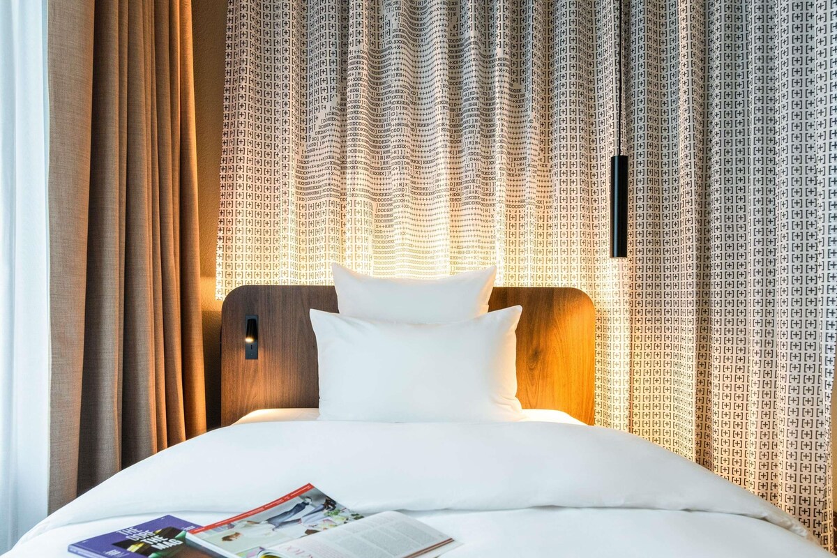 A hotel room with a neatly made bed, a magazine on the bed, and curtains with a pattern.
