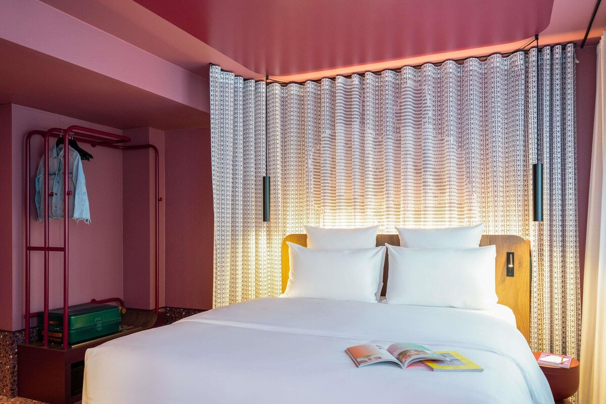 A hotel room with a bed, a nightstand, a suitcase, a coat rack, and a window with patterned curtains.