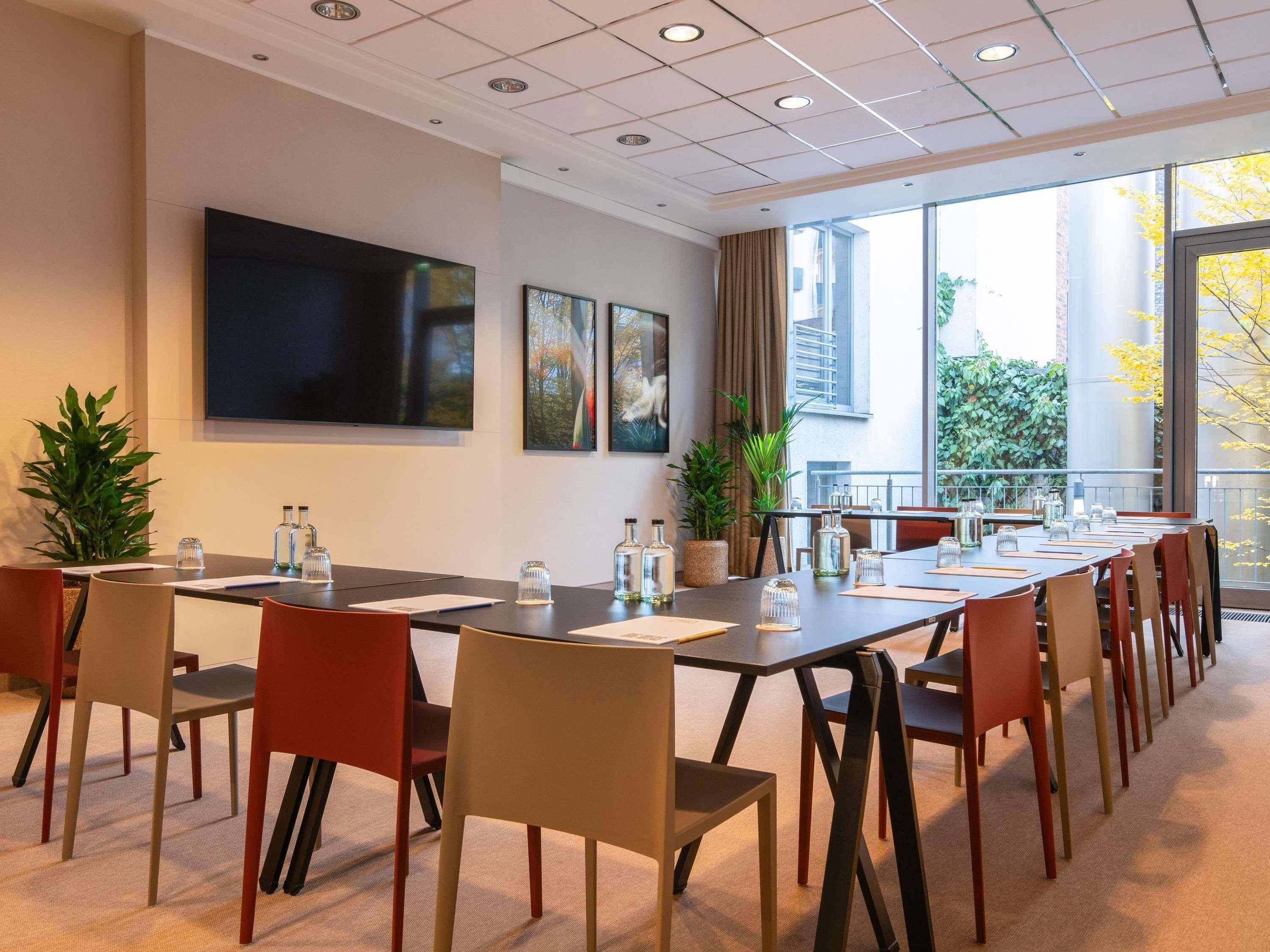 A conference room with a long table, chairs, a flat-screen TV, and large windows with a view of greenery.