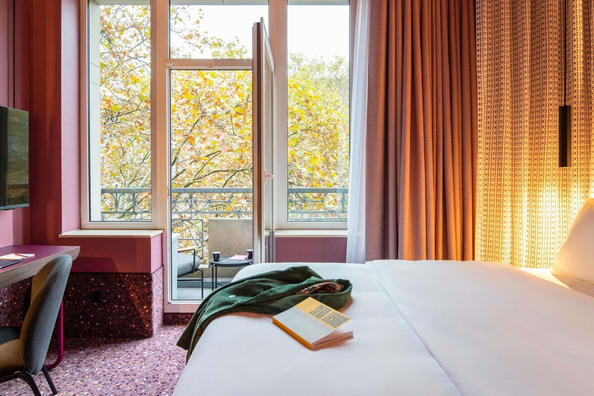 A hotel room with a large window, a bed, a desk, and a chair.