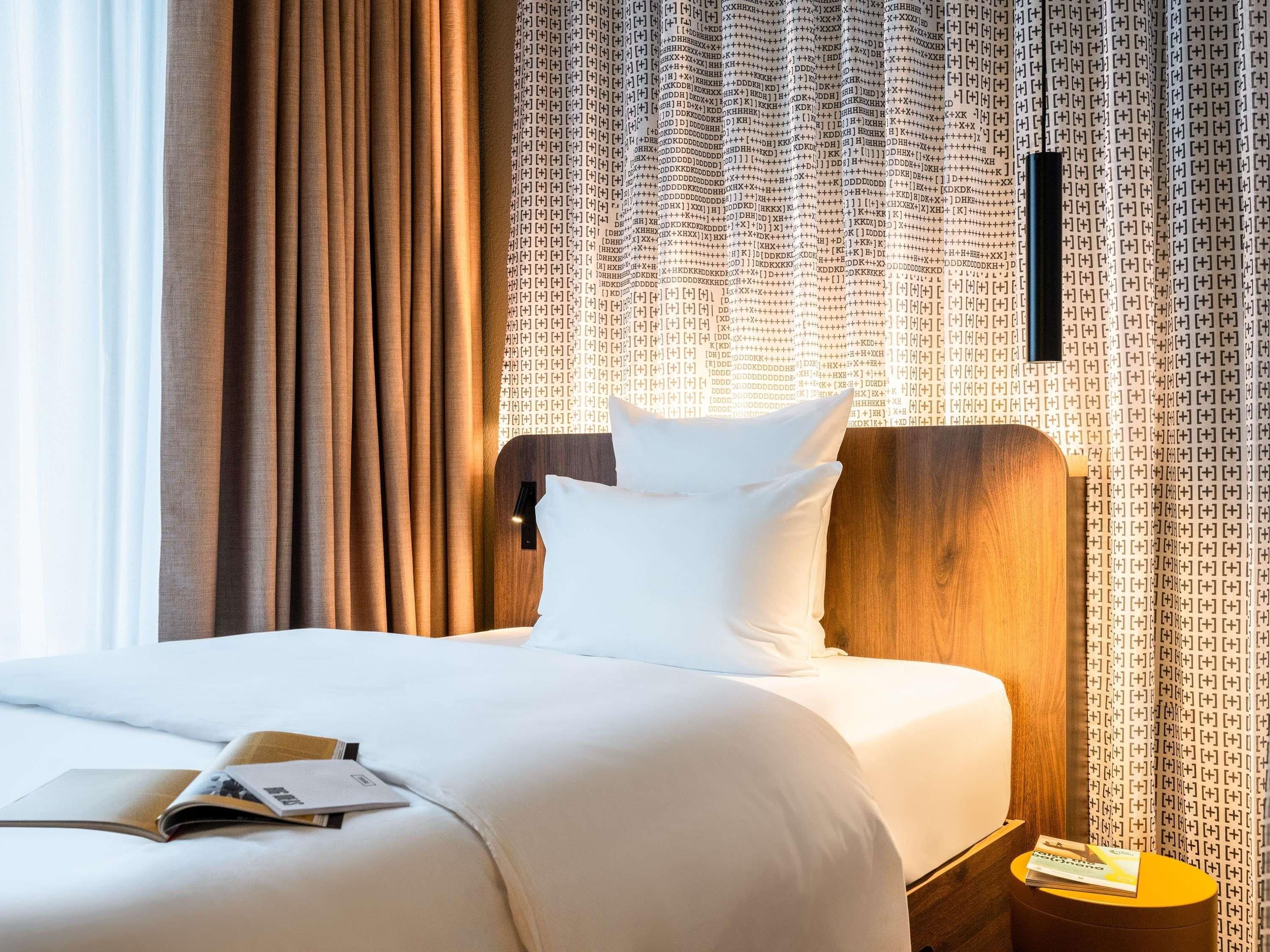 Habitación de hotel con cama, mesita de noche, lámpara y ventana con cortinas.