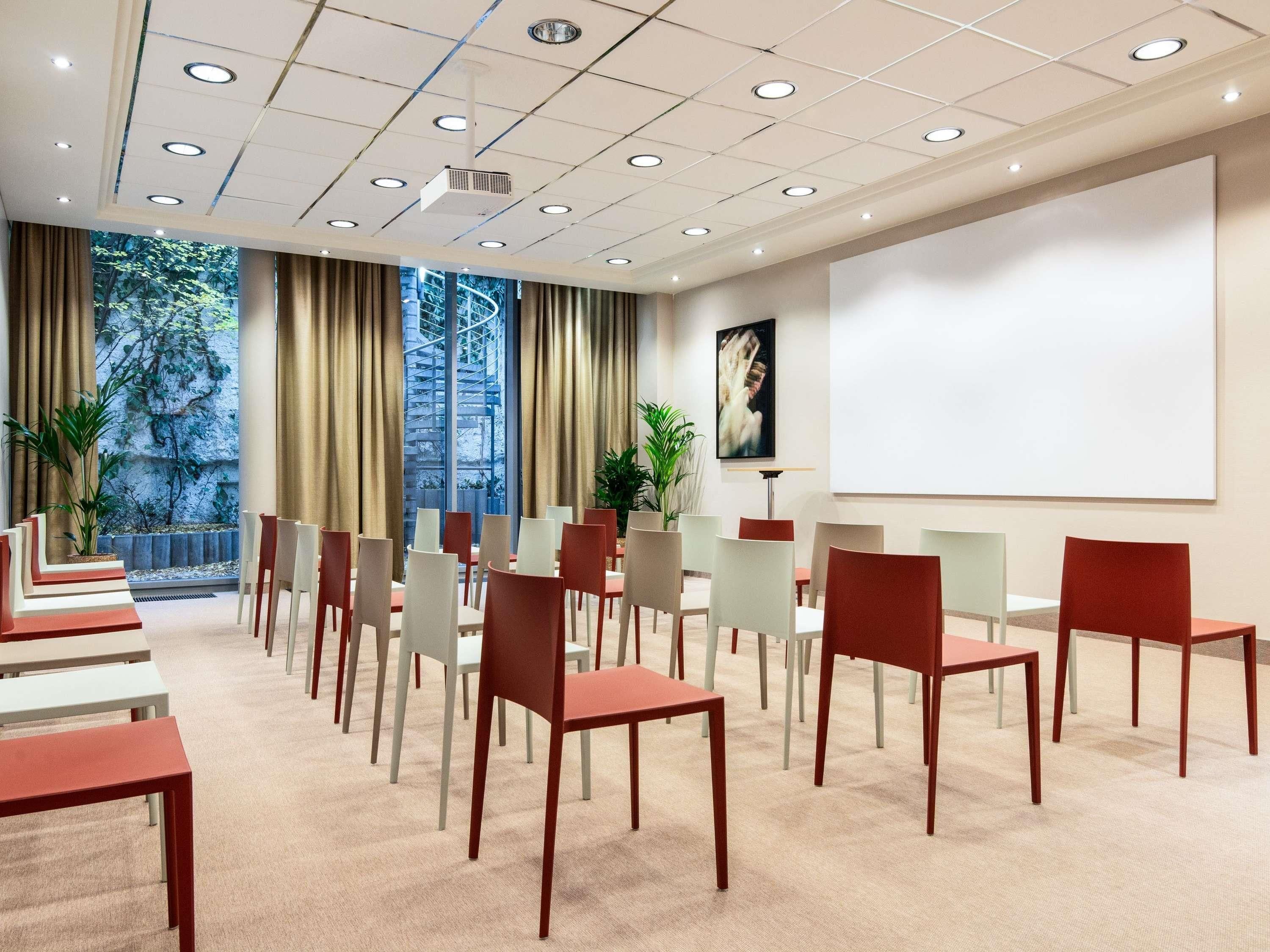 A conference room with a projector screen, multiple chairs, tables, and a large window with curtains.