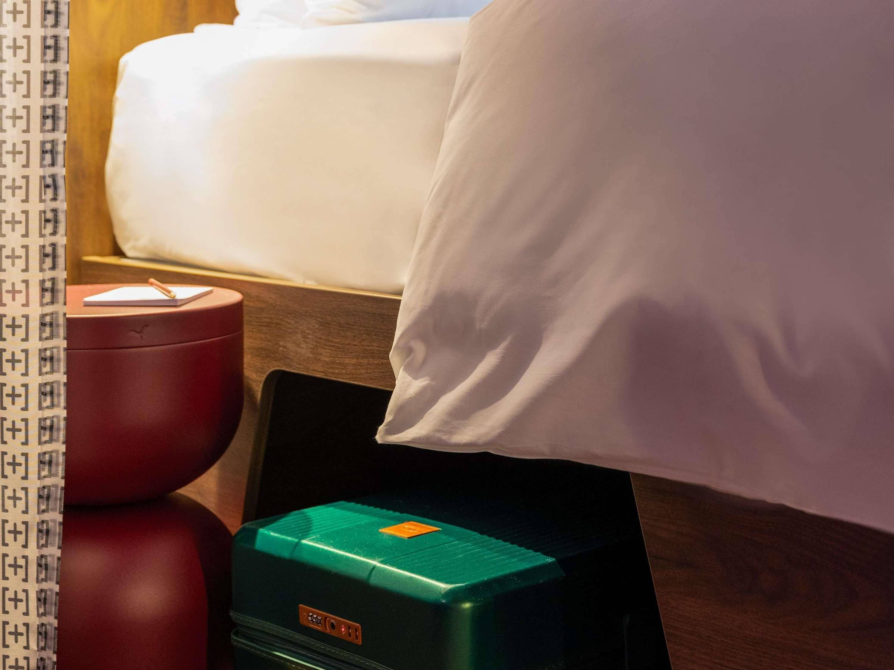 Una cama con un almohadón blanco y un objeto rojo sobre una mesita de noche de madera.