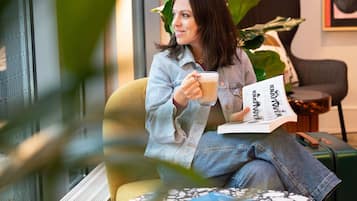 Una persona sentada en una silla amarilla, sosteniendo una taza y leyendo un libro.