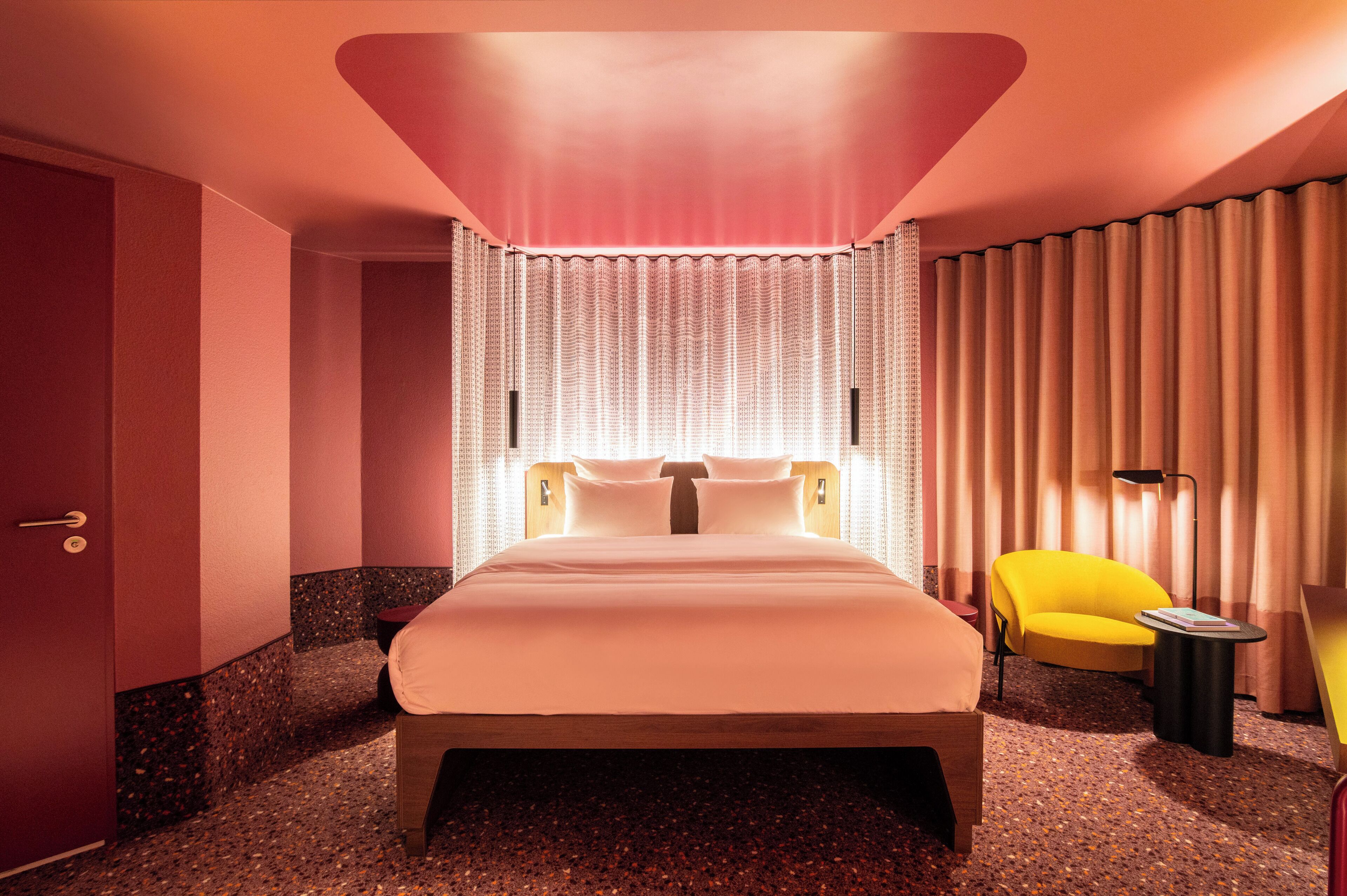 A modern hotel room with a large bed, a yellow armchair, a small side table, and a curtain-covered window.
