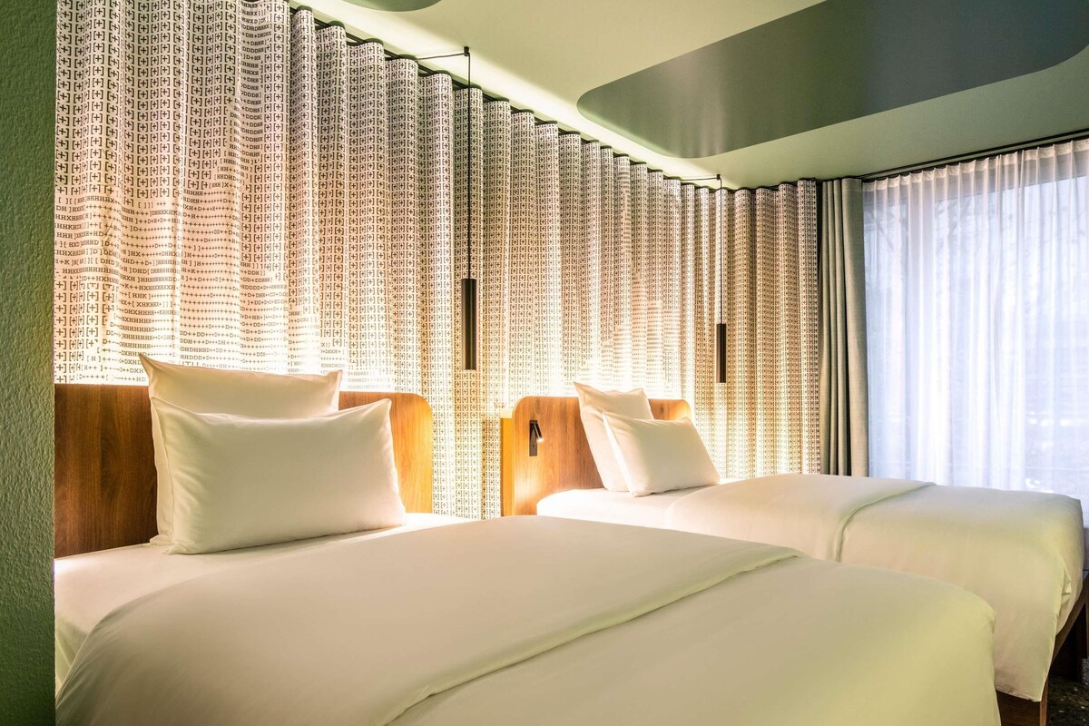 A hotel room with two beds, a large window with sheer curtains, and a wall with patterned wallpaper.