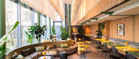 Een modern interieur met een gebogen zithoek, tafels en stoelen, omringd door grote ramen en binnenshuis planten.