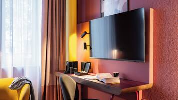 A modern hotel room with a large flat-screen TV mounted on the wall, a desk with a laptop, a coffee maker, and a chair.
