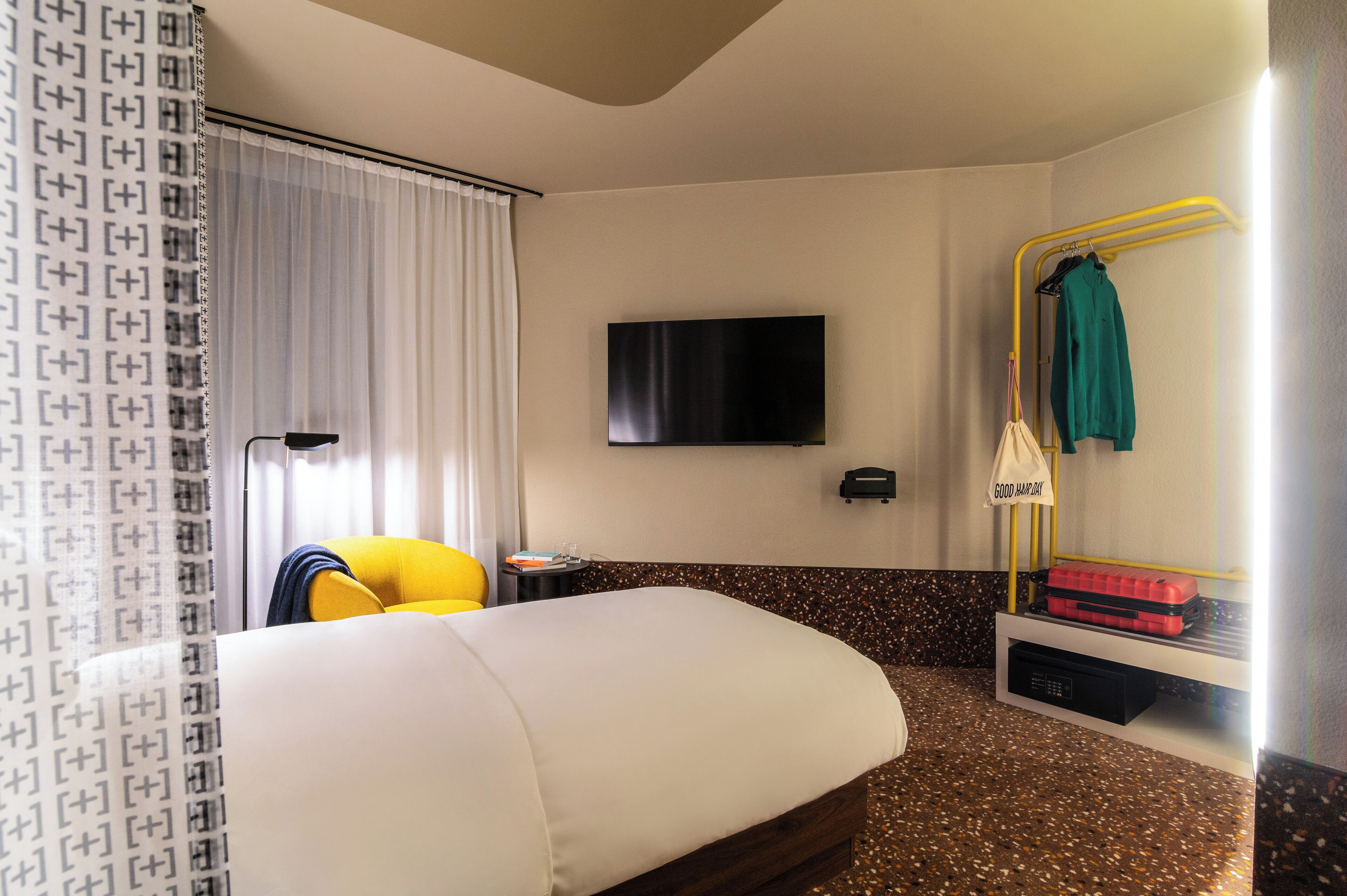 A hotel room with a bed, a TV mounted on the wall, a mirror with a towel rack, and a small table with a lamp.