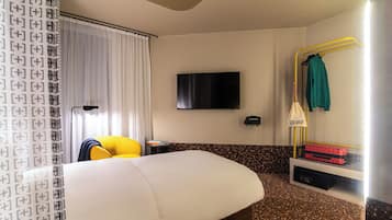 A hotel room with a bed, a TV mounted on the wall, a mirror with a towel rack, and a small table with a lamp.
