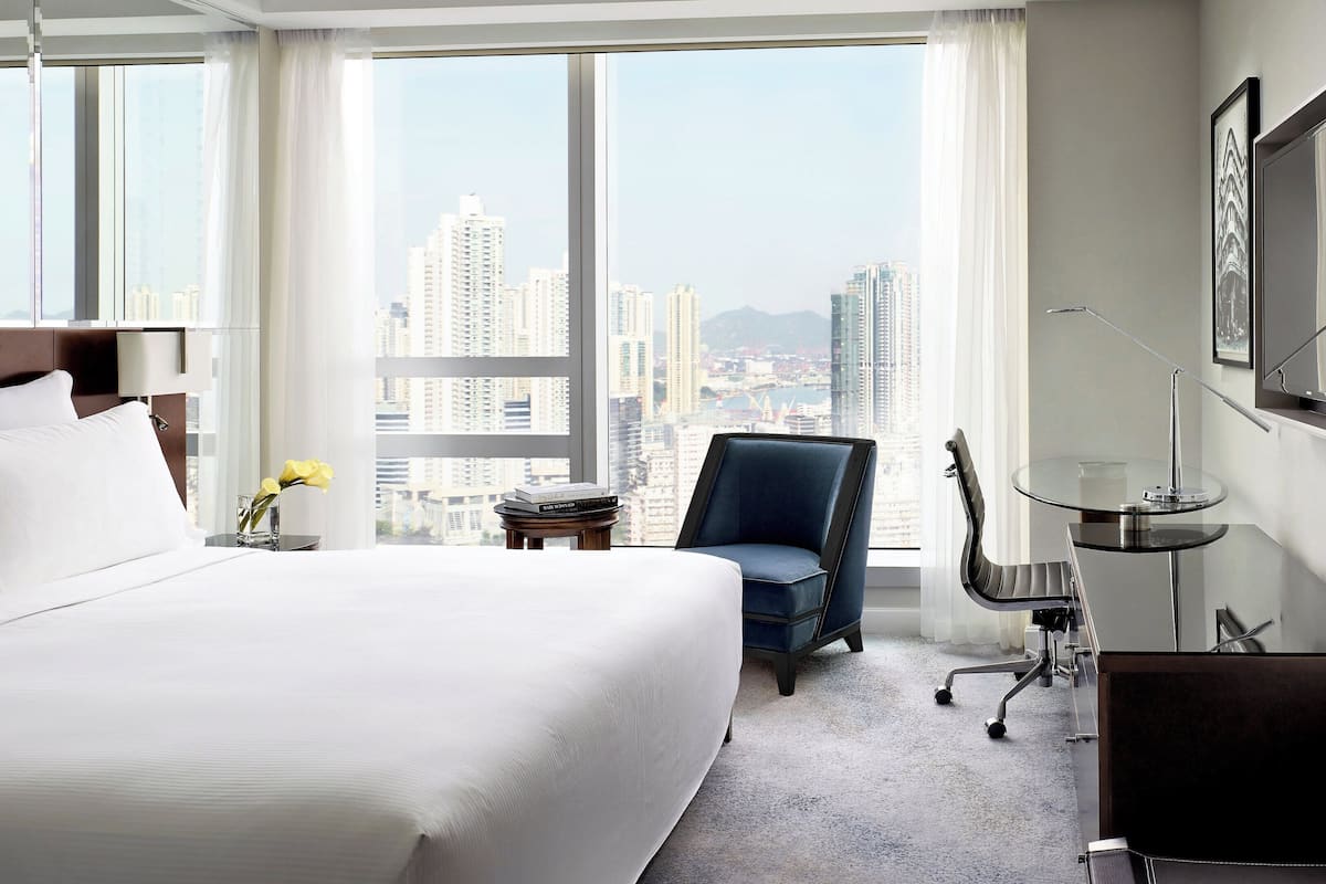 A hotel room with a large bed, a chair, a desk with a lamp, and a view of the cityscape through large windows.