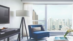 A modern hotel room with a large window offering a city view, a blue armchair, a white sofa, a flat-screen TV, and a bedside table with a lamp. - Cordis, Hong Kong (Kowloon)