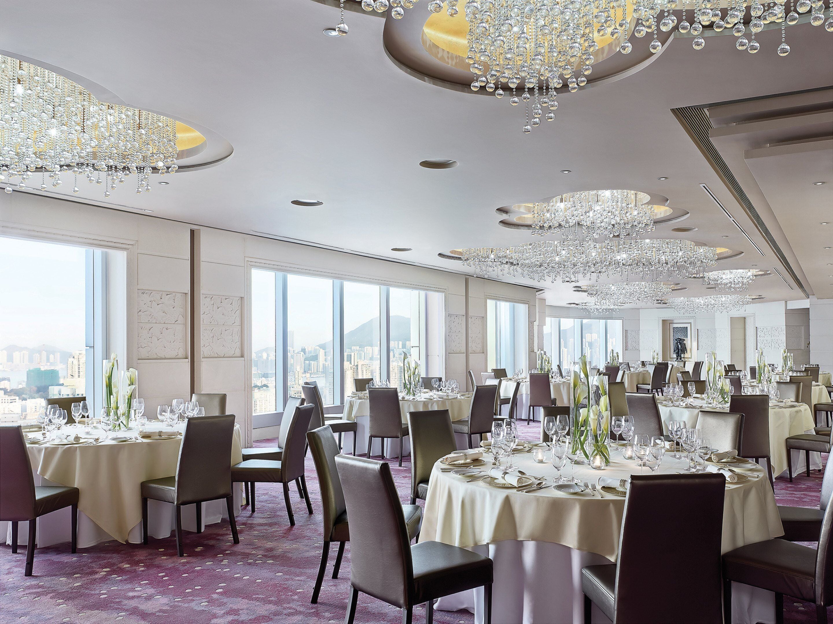 A spacious banquet hall with round tables set for dining, large windows offering a city view, and ornate chandeliers.