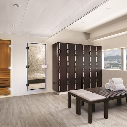 A modern bathroom with a wooden door, a mirror, a dark locker wall, and a bench with towels.
