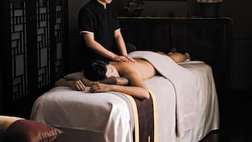 A massage therapy room with a bed, a massage table, a potted plant, and a framed artwork on the wall.