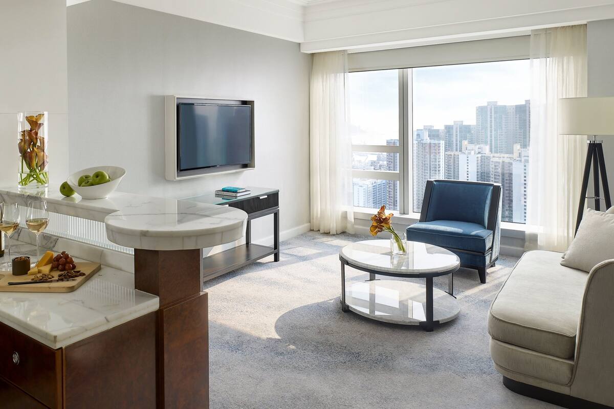 A modern living room with a marble coffee table, a sofa, a blue armchair, a flat-screen TV, and a city view through large windows.