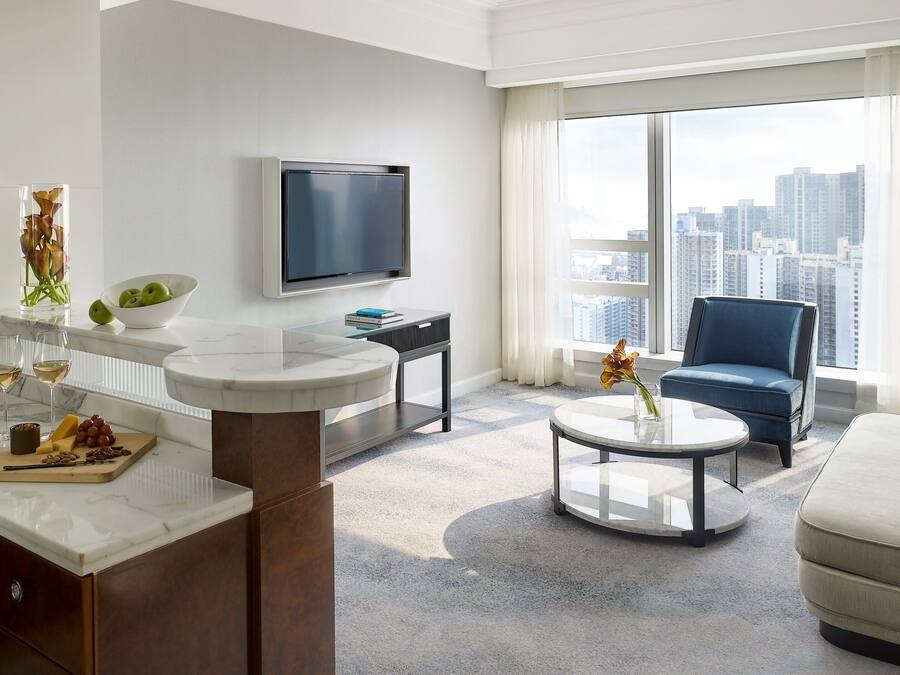 Een moderne woonkamer met een marmeren salontafel, een bank, een blauwe fauteuil, een flatscreen-televisie en een stadszicht door grote ramen.