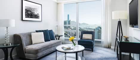 Habitación de hotel moderna con un sofá gris, un sillón azul, una mesa de centro redonda y un ventanal con vistas a la ciudad.