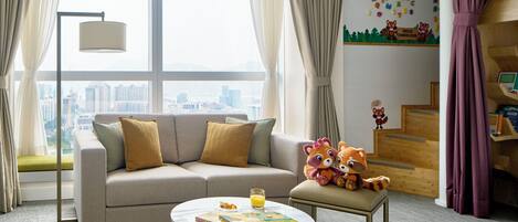 A modern hotel room with a sofa, a small table with a book and a glass of orange juice, a plush toy, and a city view through large windows.
