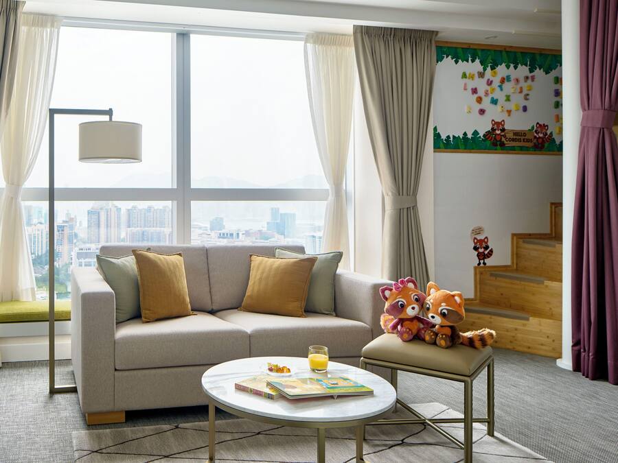 A modern hotel room with a sofa, a small table with a book and a glass of orange juice, a plush toy, and a city view through large windows.