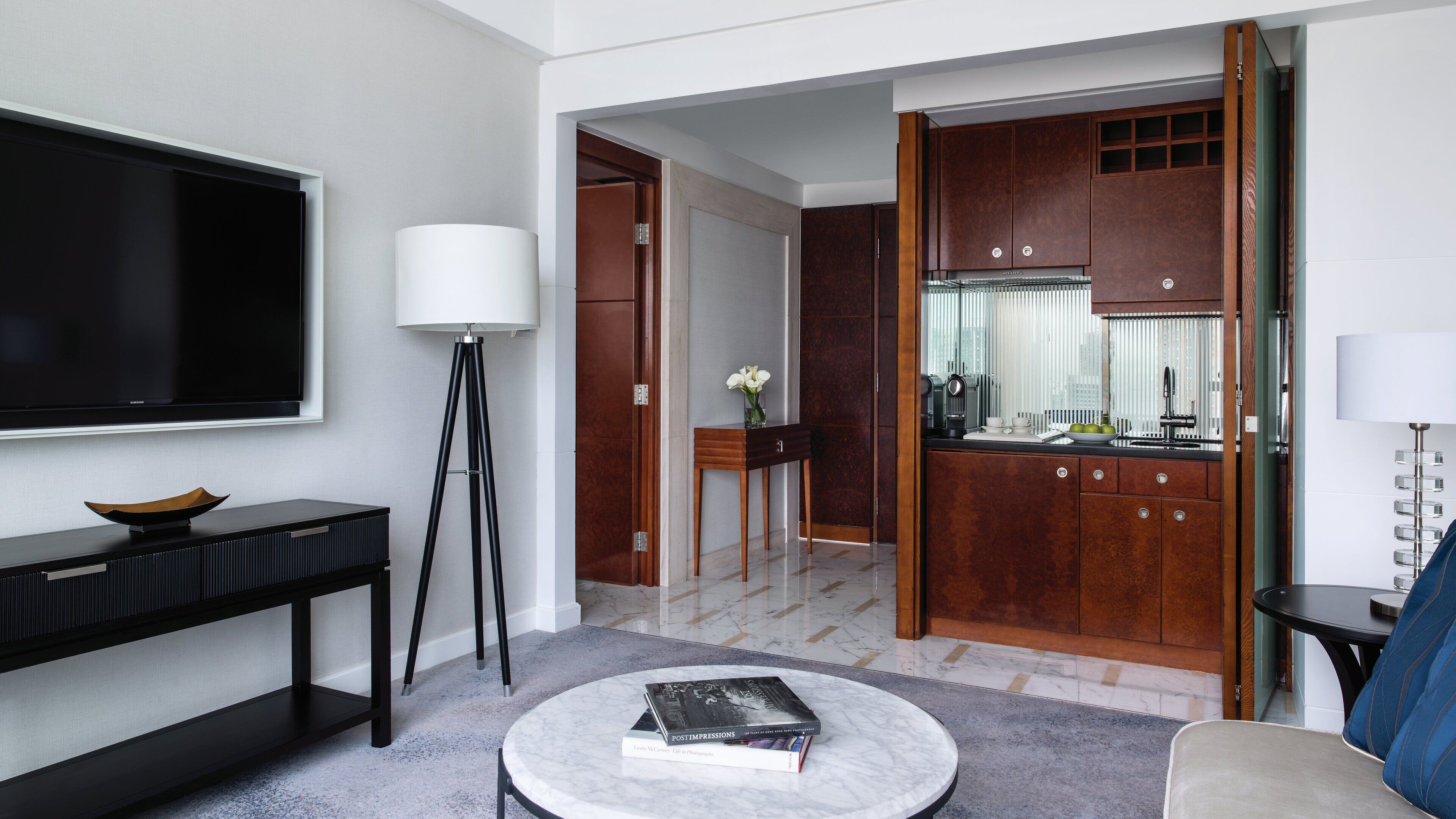 A modern hotel room with a flat-screen TV, a standing lamp, a coffee table with books, a sofa, and a view of the kitchen.