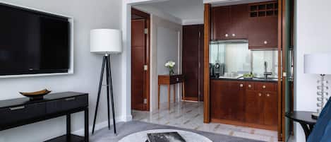 A modern hotel room with a flat-screen TV, a standing lamp, a coffee table with books, a sofa, and a view of the kitchen.
