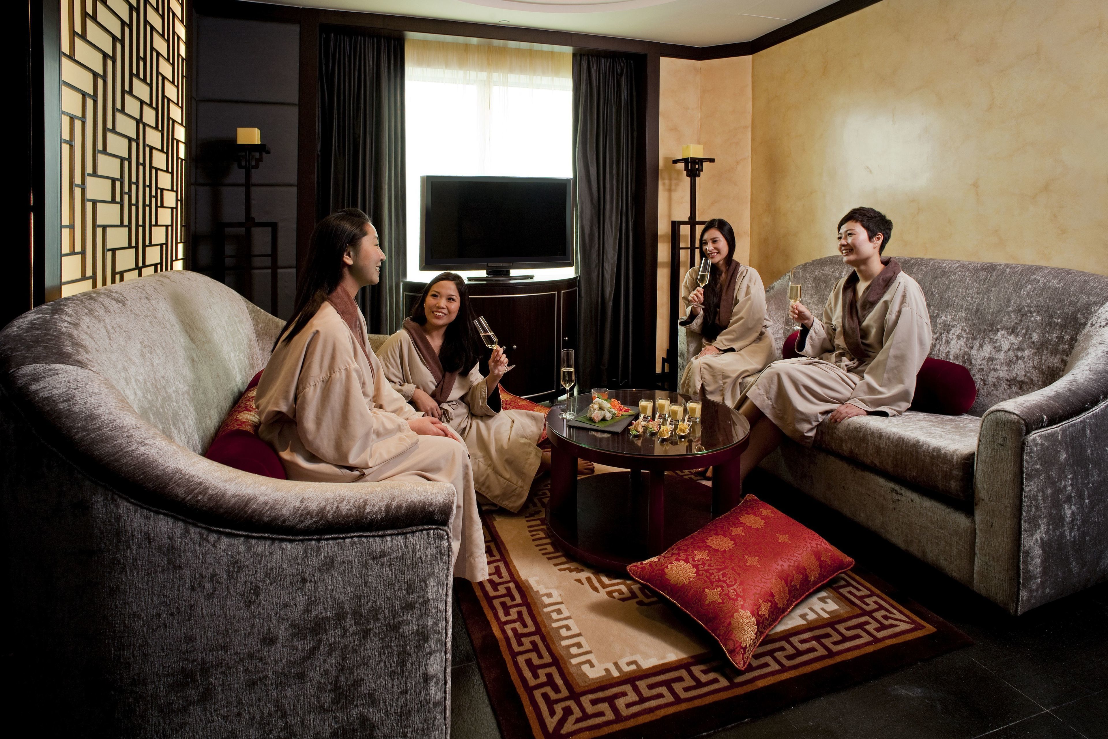 A living room with four people seated on sofas, a round coffee table with drinks, and a TV in the background.