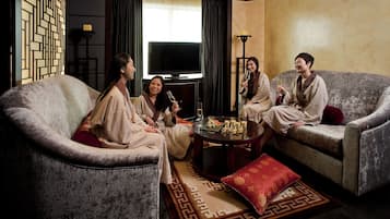 A living room with four people seated on sofas, a round coffee table with drinks, and a TV in the background.