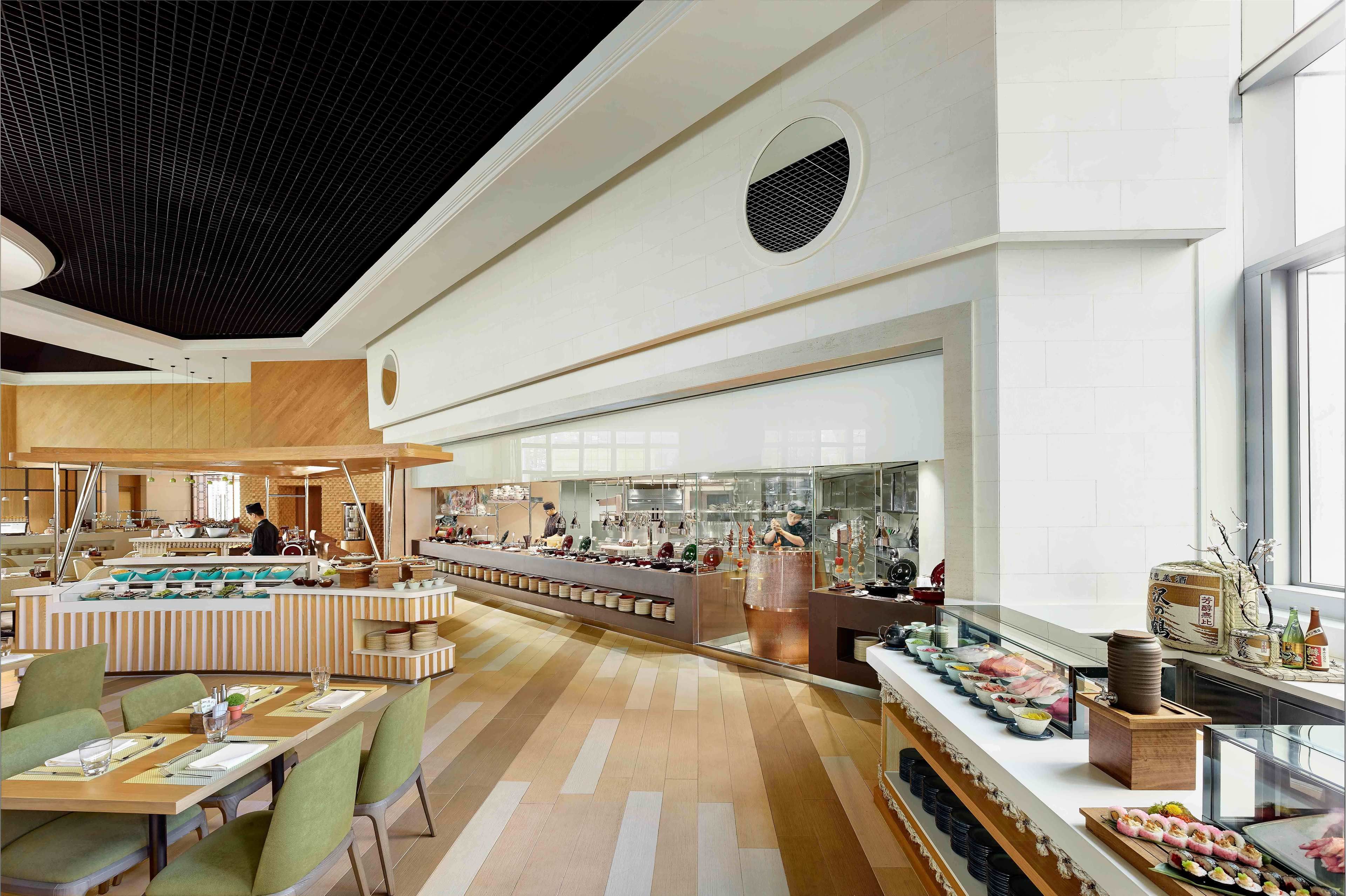 A modern restaurant with a long counter, multiple seating areas, and a kitchen visible in the background.