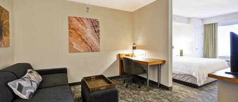 A hotel room with a sofa, ottoman, desk, chair, bed, and artwork on the wall.
