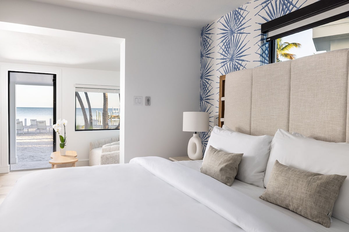 Quarto de hotel moderno com uma cama grande, vista para a praia e decoração temática tropical.