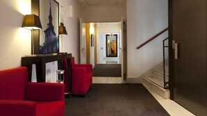 Vestíbulo de hotel con mostrador de recepción, asientos rojos, una escalera y un cuadro en la pared.