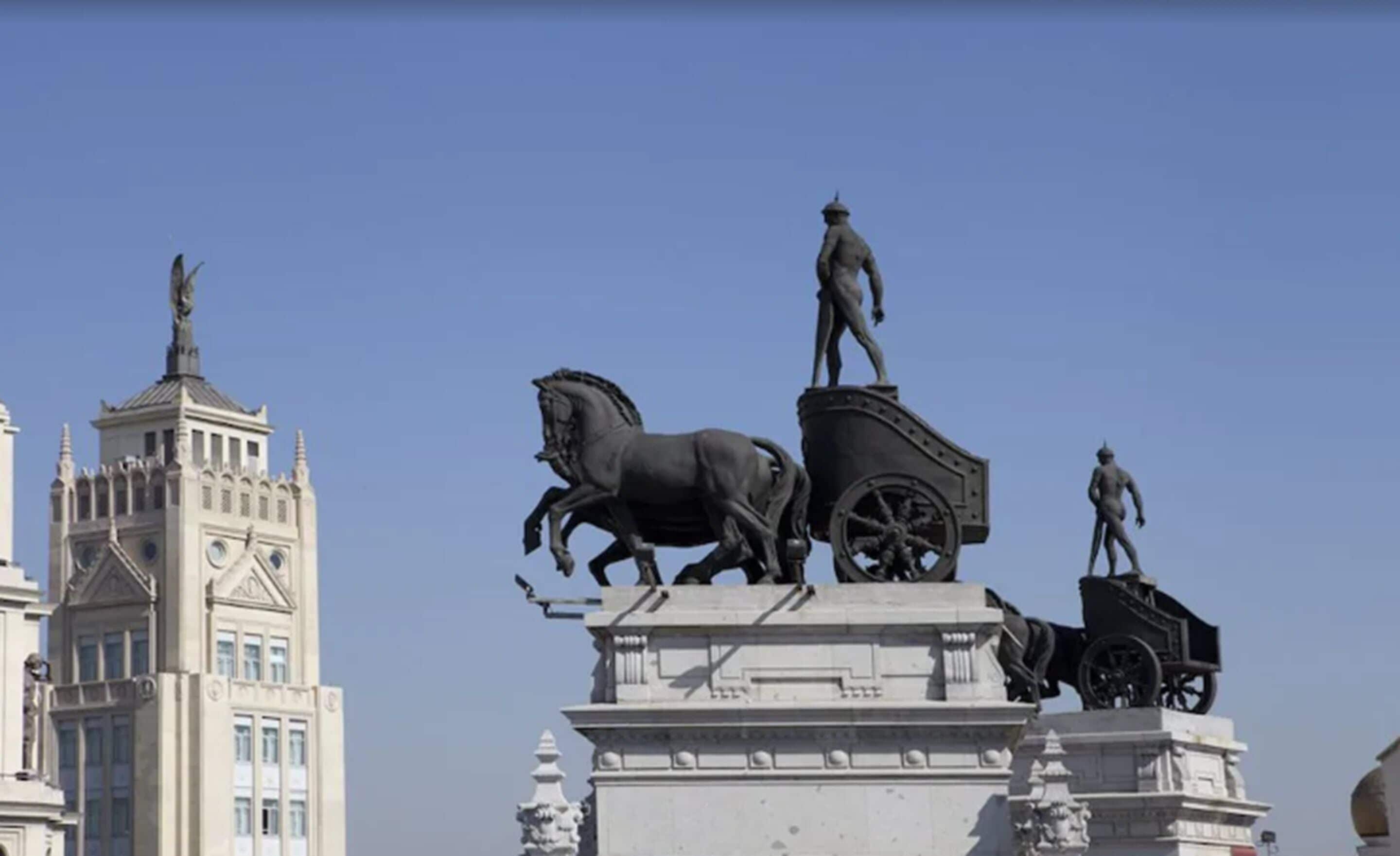 Un monumento con un carruaje tirado por caballos y dos figuras, sobre un cielo despejado.